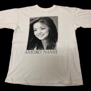 May include: White t-shirt featuring a black and white portrait of a smiling woman with long hair. The text "AMURO NAMIE" is printed below the portrait in a simple, sans-serif font. The shirt is made of a soft, light-colored fabric.