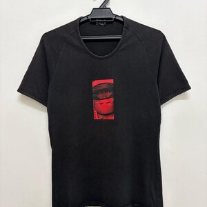 May include: Black crew neck t-shirt with a red rectangular graphic. The graphic features a face-like design with the text "YOSHIMA MATO" in red. The shirt is hanging on a black hanger.