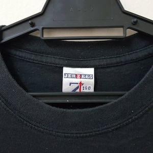 K&ouml;nnte beinhalten: Schwarzes T-Shirt mit Rundhalsausschnitt, das an einem schwarzen Kleiderb&uuml;gel h&auml;ngt. Das Etikett im Kragen lautet "JERZEES" mit einem Logo und den Zahlen "7" und "160". Das Shirt besteht aus einem weichen, dunklen Stoff.