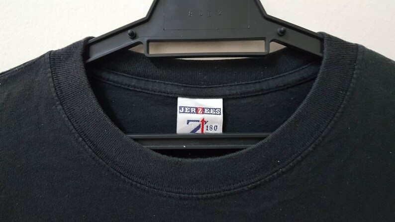 K&ouml;nnte beinhalten: Schwarzes T-Shirt mit Rundhalsausschnitt, das an einem schwarzen Kleiderb&uuml;gel h&auml;ngt. Das Etikett im Kragen lautet "JERZEES" mit einem Logo und den Zahlen "7" und "160". Das Shirt besteht aus einem weichen, dunklen Stoff.