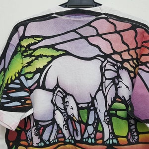 Puede incluir: Camiseta blanca con un dise&ntilde;o de vitral colorido de tres elefantes caminando por un paisaje. El dise&ntilde;o presenta tonos de morado, rosa, verde y azul.