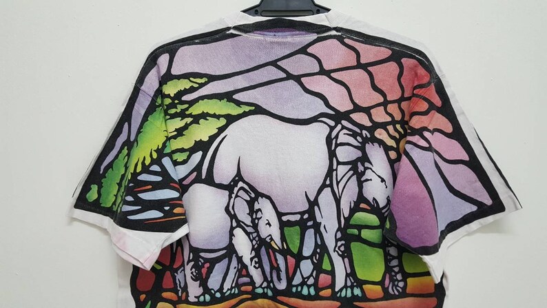 Puede incluir: Camiseta blanca con un dise&ntilde;o de vitral colorido de tres elefantes caminando por un paisaje. El dise&ntilde;o presenta tonos de morado, rosa, verde y azul.