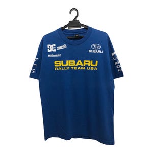 Puede incluir: Camiseta azul de Subaru Rally Team USA. La camiseta presenta el logotipo de Subaru, el texto "SUBARU RALLY TEAM USA" en amarillo y logotipos de patrocinadores. Cuelga de una percha negra.