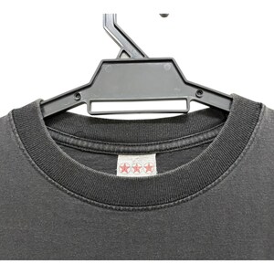 Puede incluir: Primer plano de una camiseta gris oscuro colgada de una percha de pl&aacute;stico negro. La camiseta tiene cuello redondo y una etiqueta con tres estrellas rojas. La tela parece desgastada.