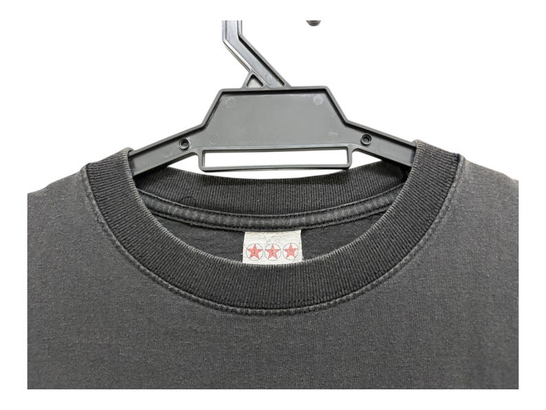 Puede incluir: Primer plano de una camiseta gris oscuro colgada de una percha de pl&aacute;stico negro. La camiseta tiene cuello redondo y una etiqueta con tres estrellas rojas. La tela parece desgastada.