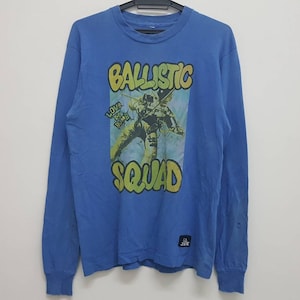 May include: A blue long-sleeved t-shirt with a graphic print of a person in a spacesuit with the text "BALLISTIC SQUAD" and "LOVE BOMB" in yellow and green lettering.