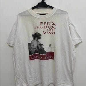 Könnte beinhalten: Weißes T-Shirt mit dem italienischen Text "Festa dell'Uva e del Vino" und "Barolino". Die Grafik zeigt eine klassische Illustration einer Person mit Trauben und einer Weinszene. Das T-Shirt hängt an einem schwarzen Kleiderbügel.