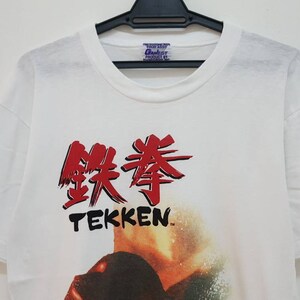 May include: White t-shirt with a red and black graphic of the Japanese characters for "Tekken" and the word "Tekken" in black below. The graphic also includes a blurry image of a person in the background.