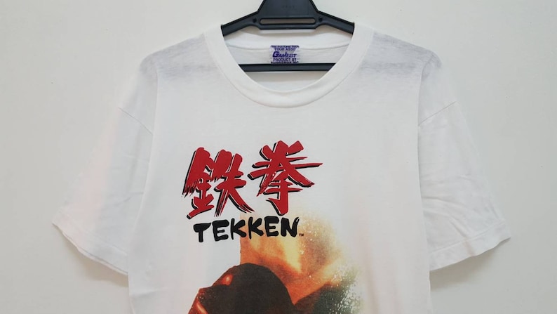 May include: White t-shirt with a red and black graphic of the Japanese characters for "Tekken" and the word "Tekken" in black below. The graphic also includes a blurry image of a person in the background.
