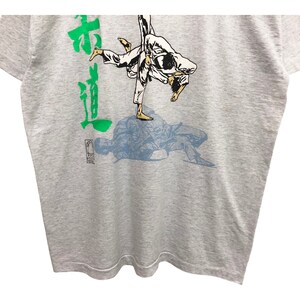 Op de afbeelding: Een grijs T-shirt met een groen Japans karakter en een blauwe en witte illustratie van twee mensen in een judowedstrijd.