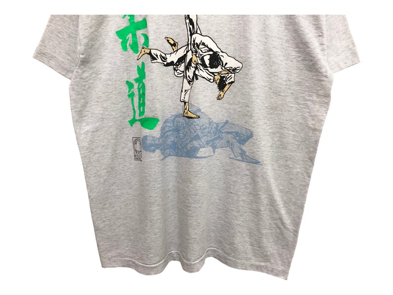 Op de afbeelding: Een grijs T-shirt met een groen Japans karakter en een blauwe en witte illustratie van twee mensen in een judowedstrijd.