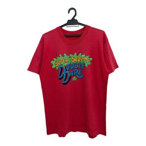 Puede incluir: Camiseta roja con el texto "Super Sloppy Double Dare" en un diseño colorido y de dibujos animados. La camiseta está colgada de una percha negra sobre un fondo blanco. El texto es en verde, azul y amarillo.