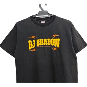 Puede incluir: Camiseta negra con el texto "DJ Shadow" en amarillo con detalles de rayos naranjas. El texto "THE OUTSIDER" est&aacute; debajo del texto principal. La camiseta est&aacute; colgada en una percha negra.