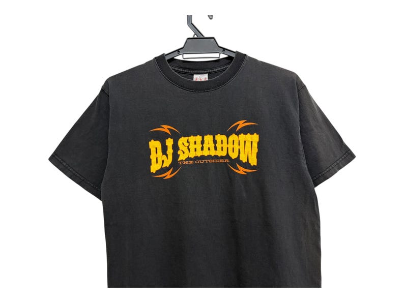Puede incluir: Camiseta negra con el texto "DJ Shadow" en amarillo con detalles de rayos naranjas. El texto "THE OUTSIDER" est&aacute; debajo del texto principal. La camiseta est&aacute; colgada en una percha negra.