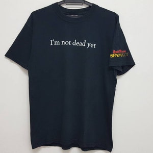 Puede incluir: Camiseta negra con el texto "I'm not dead yet" en blanco y "Monty Python's SPAMALOT" en rojo, amarillo y negro en la manga derecha.