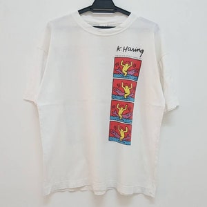 K&ouml;nnte beinhalten: Wei&szlig;es T-Shirt mit einem Grafikdruck einer gelben Figur auf rotem Hintergrund. Die Grafik ist eine Reihe von vier Bildern der Figur, die &uuml;ber ein blaues und violettes Objekt springt. Der Text "K. Haring" ist &uuml;ber der Grafik gedruckt.