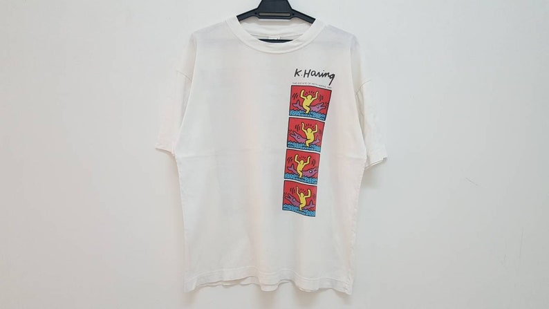 K&ouml;nnte beinhalten: Wei&szlig;es T-Shirt mit einem Grafikdruck einer gelben Figur auf rotem Hintergrund. Die Grafik ist eine Reihe von vier Bildern der Figur, die &uuml;ber ein blaues und violettes Objekt springt. Der Text "K. Haring" ist &uuml;ber der Grafik gedruckt.