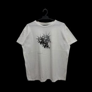 Puede incluir: Camiseta blanca con un estampado gráfico en negro y gris de una figura similar a la Estatua de la Libertad con elementos puntiagudos. La camiseta está colgada en una percha negra sobre un fondo negro.