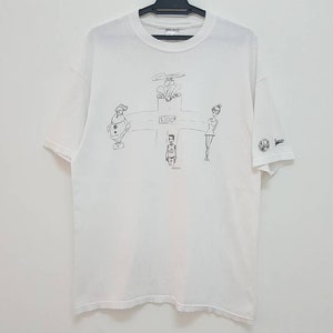 Puede incluir: Camiseta blanca con un estampado gráfico en blanco y negro de cuatro figuras. Las figuras están dibujadas en un estilo de dibujos animados y están de pie en formación de cruz. El texto "B.L.O.O.D." está impreso en el centro del gráfico.