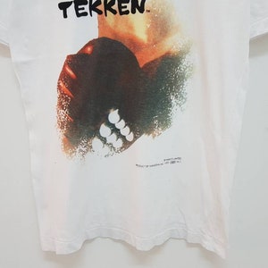 May include: White t-shirt with a graphic print of a character from the video game "Tekken". The character is depicted in a stylized, abstract style with a red, orange, and green color scheme.