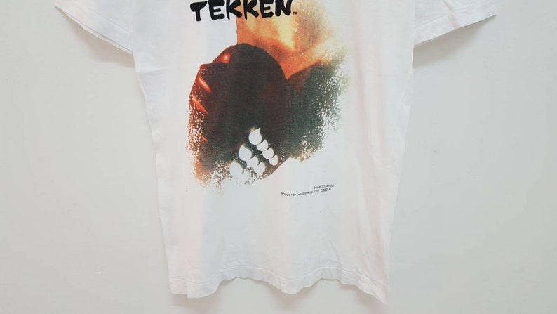May include: White t-shirt with a graphic print of a character from the video game "Tekken". The character is depicted in a stylized, abstract style with a red, orange, and green color scheme.