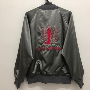 Puede incluir: Una chaqueta bomber de satén gris con una telaraña bordada en rojo y una figura en el centro. El texto "Kiss of the Spider Woman" está bordado debajo de la telaraña.