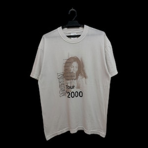 Op de afbeelding: Lichtbeige t-shirt met een afbeelding van een vrouw en de tekst "Magnum Tour 2000". Het shirt hangt aan een zwarte hanger tegen een zwarte achtergrond.
