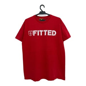 Puede incluir: Una camiseta roja con la palabra "FITTED" en letras blancas en el pecho. La palabra "FITTED" está encima de un emblema en forma de escudo. La camiseta está colgada de una percha negra sobre un fondo blanco.