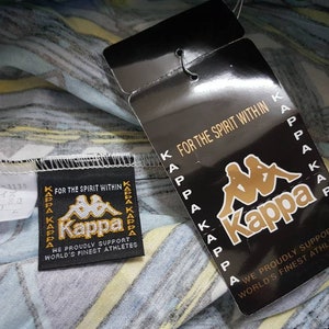 May include: A white, blue, and yellow patterned fabric with a black and white Kappa logo tag. The tag reads "For the spirit within" and "We proudly support world's finest athletes".