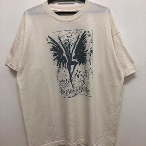 May include: Cream-coloured t-shirt featuring a dark grey graphic of a winged figure and the text "So I Had To Sell Him". The graphic is in a rectangular frame with a distressed texture. The shirt is hanging on a black hanger.