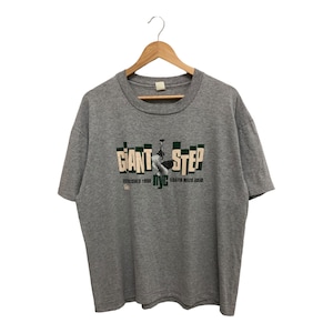 May include: A gray t-shirt with a graphic print of a person playing a saxophone. The text "Giant Step" is printed in green and white letters.