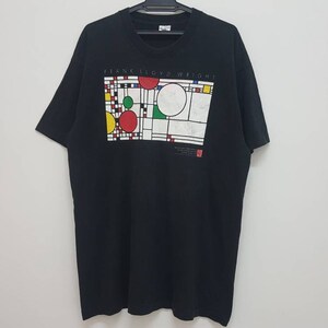 Op de afbeelding: Zwart T-shirt met een grafische print van een geometrisch ontwerp in rood, geel, groen en blauw. De tekst "Frank Lloyd Wright" is boven het ontwerp gedrukt.