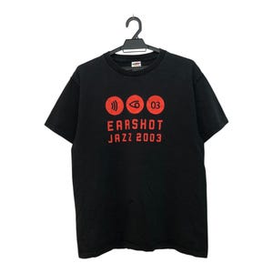 May include: Black t-shirt with a red graphic that reads "EARSHOT JAZZ 2003". The graphic includes three red circular icons. The t-shirt is hanging on a black hanger.