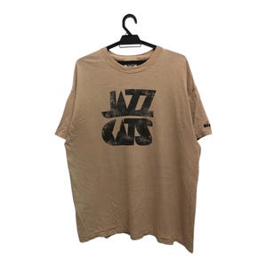 May include: Tan t-shirt with the text "Jazz Cats" in a distressed black font. The shirt is hanging on a black hanger against a white background. The shirt has short sleeves and a crew neck.