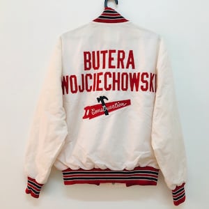 May include: White bomber jacket with red and black striped cuffs and collar. The back of the jacket features red embroidered text that reads "BUTERA WOJCIECHOWSKI" and a red and black illustration of a hammer and a banner that reads "Construction".