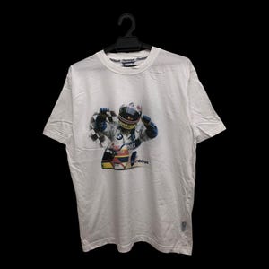 May include: White t-shirt featuring a colorful graphic of a race car driver in a helmet holding a checkered flag. The design includes the BMW logo and other racing details. The shirt is hanging on a black hanger.