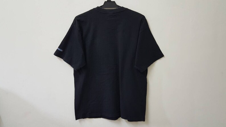 K&ouml;nnte beinhalten: Ein schwarzes T-Shirt mit kurzen &Auml;rmeln, das an einem schwarzen Kleiderb&uuml;gel h&auml;ngt. Die R&uuml;ckseite des T-Shirts ist sichtbar, mit einem kleinen Textdetail am linken &Auml;rmel. Das T-Shirt ist aus einem weichen Material.