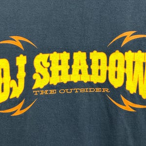 Puede incluir: Camiseta azul oscuro con el texto amarillo "DJ SHADOW" y las palabras "THE OUTSIDER" debajo. Dise&ntilde;os de rayos naranjas est&aacute;n encima y debajo del texto.
