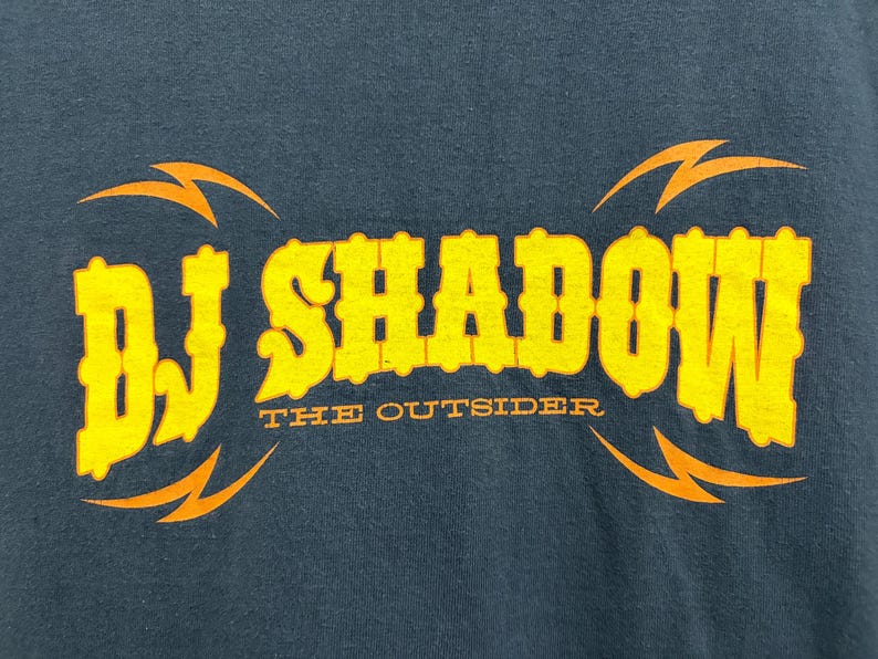 Puede incluir: Camiseta azul oscuro con el texto amarillo "DJ SHADOW" y las palabras "THE OUTSIDER" debajo. Dise&ntilde;os de rayos naranjas est&aacute;n encima y debajo del texto.
