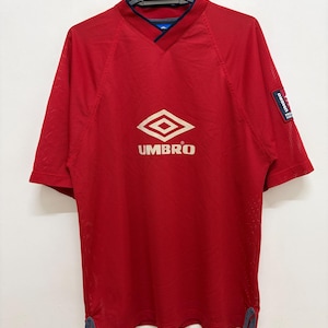 May include: Red short-sleeved athletic jersey with a V-neck collar and the Umbro logo in the center. The jersey has a mesh-like texture and a small Umbro patch on the right sleeve. The jersey is hanging on a black hanger.