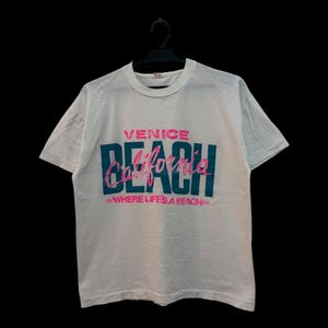 May include: White t-shirt with the words "Venice Beach California" in teal and pink lettering. The text "Where life's a beach" is below the main text. The shirt is hanging on a black hanger.