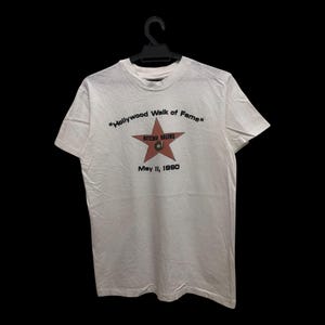 May include: White t-shirt featuring a red star graphic with the text "Hollywood Walk of Fame" and "Ritchie Valens" in black. The date "May 11, 1990" is printed below the star. The shirt is hanging on a black hanger.