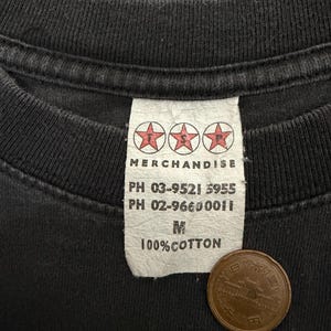 Puede incluir: Primer plano de una camiseta negra con una etiqueta blanca. La etiqueta dice "MERCHANDISE" encima de tres logotipos de estrellas rojas. Debajo, dos n&uacute;meros de tel&eacute;fono, la talla "M" y "100% COTTON". Una moneda japonesa est&aacute; sobre la camiseta.