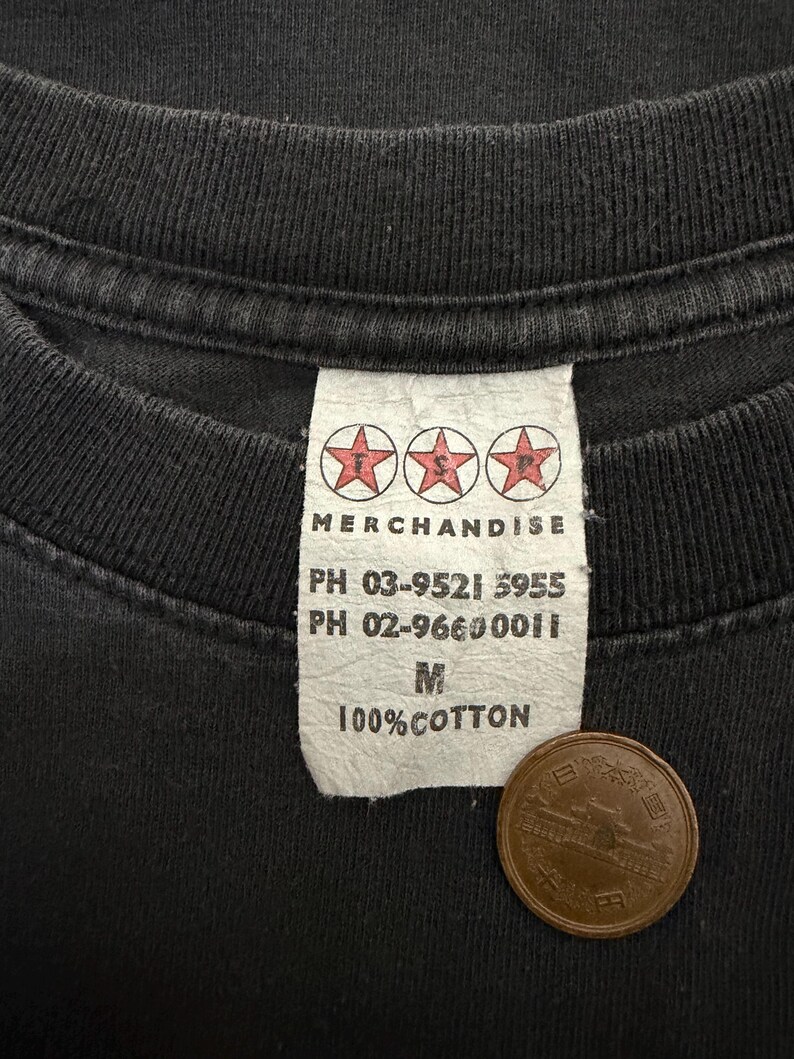 Puede incluir: Primer plano de una camiseta negra con una etiqueta blanca. La etiqueta dice "MERCHANDISE" encima de tres logotipos de estrellas rojas. Debajo, dos n&uacute;meros de tel&eacute;fono, la talla "M" y "100% COTTON". Una moneda japonesa est&aacute; sobre la camiseta.