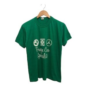 Op de afbeelding: Groen T-shirt met korte mouwen met witte geborduurde logo's van BMW, Rover en Mercedes-Benz. De tekst "Foreign Car Specialists" is ook in wit geborduurd.