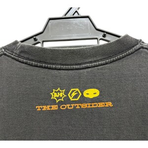 Puede incluir: Camiseta gris oscuro con cuello redondo, colgada de una percha negra. La camiseta presenta gr&aacute;ficos amarillos y naranjas, incluyendo un rayo y una cara, con el texto "THE OUTSIDER" debajo.