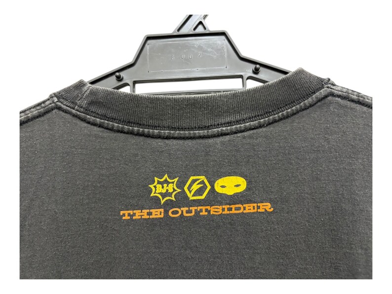 Puede incluir: Camiseta gris oscuro con cuello redondo, colgada de una percha negra. La camiseta presenta gr&aacute;ficos amarillos y naranjas, incluyendo un rayo y una cara, con el texto "THE OUTSIDER" debajo.