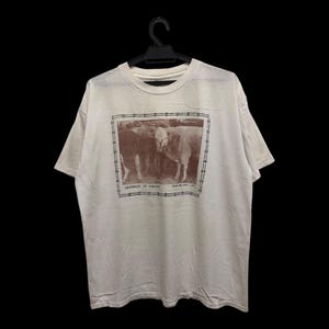 May include: Off-white t-shirt featuring a sepia-toned image of two cows within a barbed wire frame. The text "I'm surrounded by idiots" is printed below the image. The shirt is hanging on a black hanger.