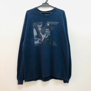 May include: A navy blue long-sleeved t-shirt featuring a faded graphic print of a man holding a gun. The graphic is in the shape of a letter "K".