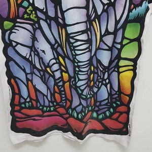 Puede incluir: Una camiseta blanca con un dise&ntilde;o de vitral colorido de tres elefantes. Los elefantes est&aacute;n delineados en negro y rellenos con tonos de azul, morado, verde, amarillo y naranja.